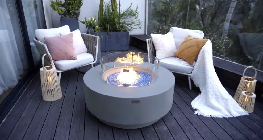 Contemporary outdoor fire table with blue glass pieces and a flame, with comfortable seating and plants in the background on a wooden deck.