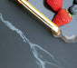 A close-up shot featuring two strawberries, a blueberry, and a gold and silver utensil resting on a dark gray, marble-patterned surface.