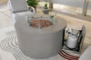 Light gray round fire pit table with lava rocks, glass wind guard, and visible propane tank, set on a patterned rug indoors.