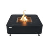 A modern, square fire pit table with a black marble-look top and a stainless steel burner is lit with a flickering flame. The table has a black base and is elevated on small silver feet.