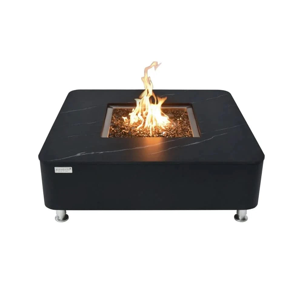 A modern, square fire pit table with a black marble-look top and a stainless steel burner is lit with a flickering flame. The table has a black base and is elevated on small silver feet.