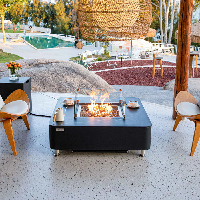 A modern, square gas fire table with a black finish and a glass wind guard. The table is surrounded by comfortable seating and features a flame that dances within the fire pit. The backdrop is a beautiful outdoor space with large rocks and greenery.