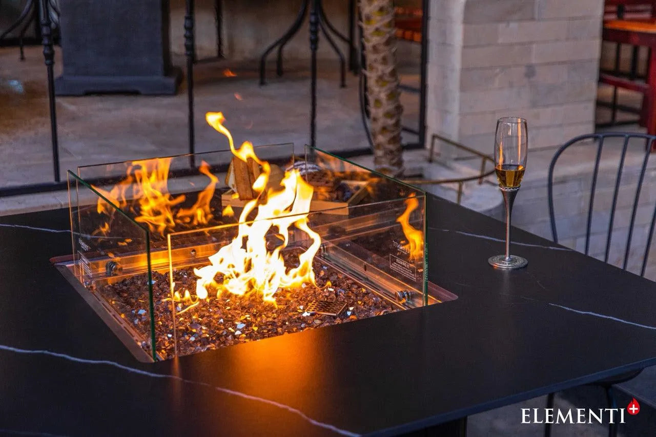  Close-up of a square black fire pit table with a glass surround containing a lit fire. A champagne flute sits on the table's edge. The "ELEMENTI+" logo is visible in the bottom right.