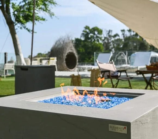 elementi light grey square pit table lit on blue fire glass with propane square fire tank next to it in a backyard with outdoor furniture laid out