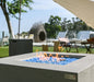 elementi light grey square pit table lit on blue fire glass with propane square fire tank next to it in a backyard with outdoor furniture laid out