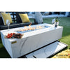 A sleek, rectangular fire table with a white marble-patterned porcelain top is the central focus on a green lawn. Flames dance above a bed of blue glass within the recessed center of the table. 