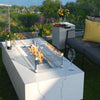 A rectangular fire pit table with a glass enclosure. The fire pit is burning with a small flame and is filled with blue fire glass. There is a wine bottle and two wine glasses on the table. There is a small side table next to the fire pit table with a yellow throw pillow on it. There is a couch behind the fire pit table with a yellow throw pillow on it.