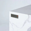 A close-up of the Elementi logo on the side of a white marble porcelain fire pit table. The logo features the word "Elementi" in a bold font, with the tagline "The Art of Living" positioned below it.
