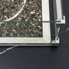 Close-up of reflective fire glass in a stainless steel burner with a glass wind guard on an Elementi fire pit table. The Elementi logo is visible.