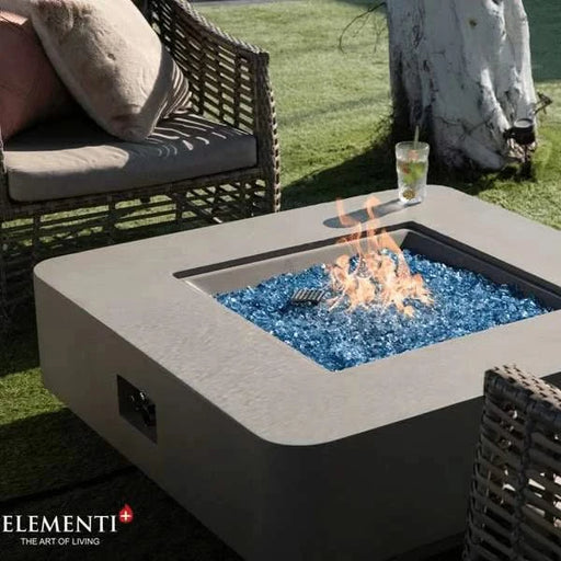 square fire pit table with blue fire glass burning and cold glass of drink is on the side of the table. fire table is on top of grass in an outdoor setting