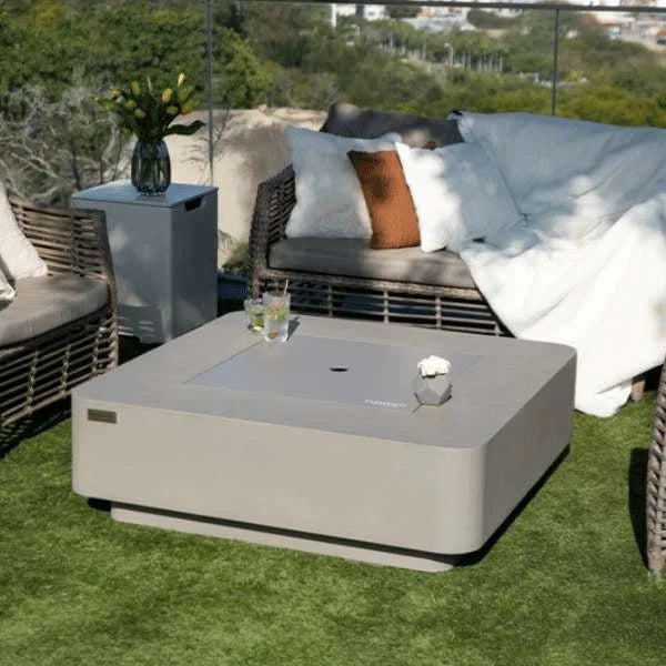 elementi plus square fire pit table with fire pit lid on grass in outdoor patio. there is cold drinks placed on top of the fire table. there is a sofa with cushions and a matching tank cover to the fire table that is being ased as a table top to hold a vase of flowers