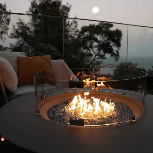 round fire pit table lit on blue fire glass in the evening in patio in the middle of outdoor furniture. round windscreen is around burning fire