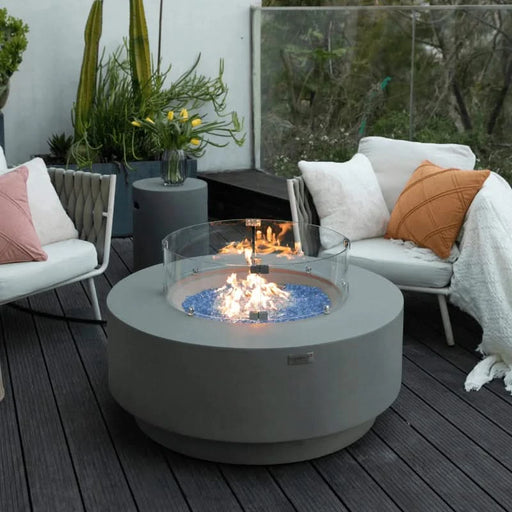 light grey round fire pit table with round wind screen. fire pit is lit on blue fire glass with furniture around it