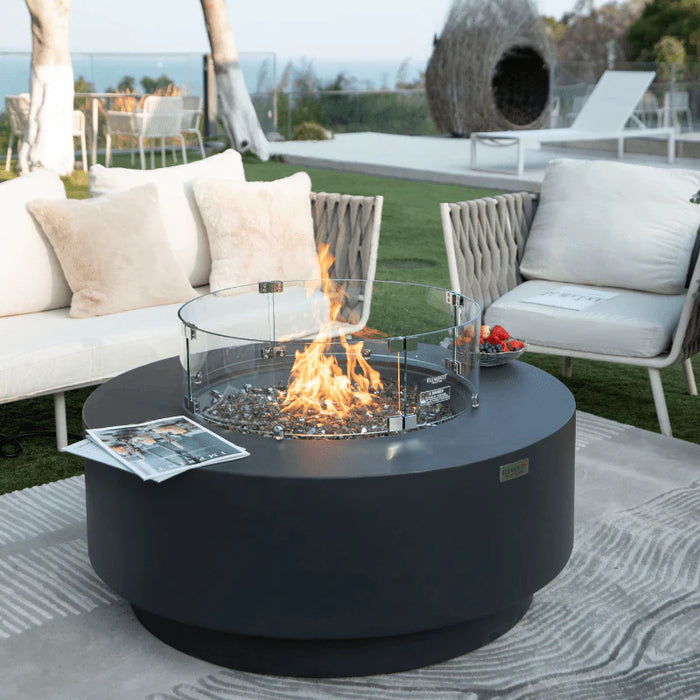 fire pit table round outside in backyard. round fire pit is burning yellow and orange flames on fire glass. fire flames is protected by round glass windscreen. there is a magazine and fruit platter on round fire table