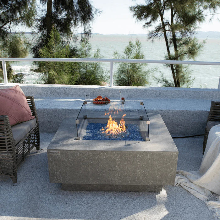 light grey textured square fire pit table with glass wind screen. fire pit is lit with fruit and glass on the side