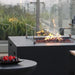 Dark gray fire pit table with flames, lava rocks, and a glass wind guard on an outdoor patio, with a bowl of fruit in the foreground.