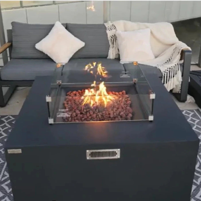 Dark gray fire pit table with flames, lava rocks, and a glass wind guard, set in front of a gray sofa on a patio.