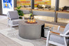 Gray, round Fire Table with flames burning among lava rocks, positioned on a patterned rug in a modern indoor or covered patio setting with gray armchairs and a built-in seating area in the background. A propane tank is visible through a side opening.