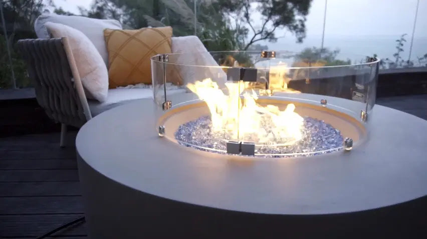 Image of a round fire pit with flames and a curved glass wind guard, with patio furniture and a view in the background.