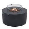 Modern, round fire pit table in dark gray with flames burning among lava rocks, surrounded by a clear glass wind guard with metal fasteners.