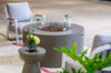 Modeno Tramore or York fire table with lava rocks and glass wind guard, in a patio setting with modern furniture and plants.