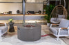 Gray Modeno Tramore Fire Table with lava rocks and a glass wind guard, set on a patio with gray armchairs, a side table with flowers, a hanging chair, and plants.