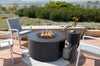 Dark gray round fire pit table with flames, surrounded by patio furniture, showing a propane tank through a side opening.