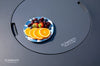Dark gray fire pit table lid with a plate of sliced oranges, strawberries, blueberries, and cherries. The "ELEMENTI" brand logo is shown.