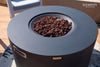 Dark gray, round fire pit table with brown lava rocks in the center. The "ELEMENTI THE ART OF LIVING" brand logo is visible in the upper right corner, and a smaller "ELEMENTI" label is on the side of the table.