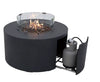 Modern, round fire pit table in dark gray with a clear glass wind guard surrounding the flames and a visible propane tank compartment with a sliding door.