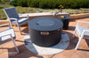 Dark gray round fire pit table with lid, surrounded by patio furniture, with the "ELEMENTI" brand logo visible.