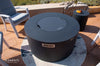 Dark gray round fire pit table with lid, surrounded by patio furniture, with the "ELEMENTI" brand logo visible.