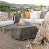 A large, round, concrete fire pit with a textured, rock-like finish sits on a wooden deck. The fire pit has a central bowl with a bright orange flame burning inside. A glass of wine sits on the edge of the fire pit, and there are pillows and a blanket nearby. The scene suggests a cozy outdoor gathering space.