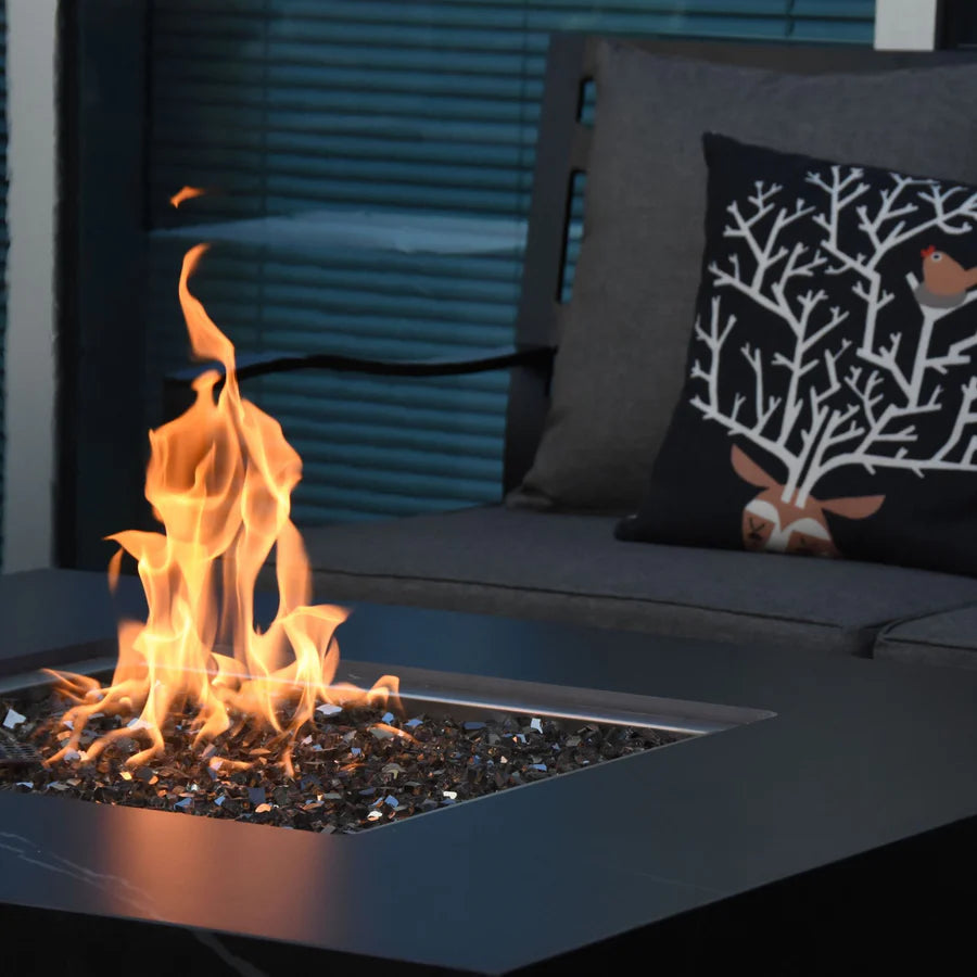 Close-up of a square fire pit with a strong flame burning in the center, with a dark gray couch and a decorative pillow in the background.