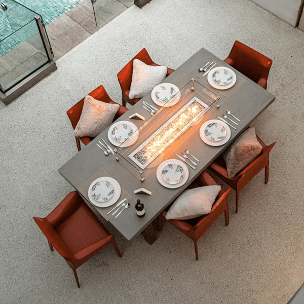 top view of outdoor dining set with fire pit in middle. fire pit is lit and rectangle glass windshield over it. there are plate, spoons and forks laid out on table