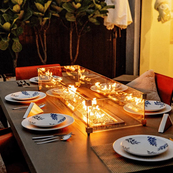 outdoor dining table with fire pit and dining table is set with plates, knife and spoon. fire fit is lit on fire glass and yellow and orange flames are blowing. rectangle windscreen is over fire pit