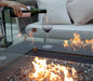 lady pouring wine in wine glass that is on fire pit table that is lit on fire glass. they are outside on a patio with the fire pit table in middle of outdoor furniture