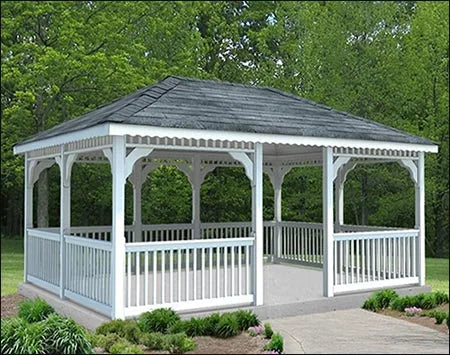 White vinyl rectangle gazebo with a double roof. The gazebo has a rectangular shape with a gabled roof and white railings. The gazebo is surrounded by green trees and grass.