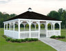 The image shows a white vinyl rectangular gazebo with a double roof and a cupola. The gazebo features a gabled roof, white railings, and a dark-colored roof. The gazebo is surrounded by green trees and grass. There is a paved walkway leading up to the gazebo.