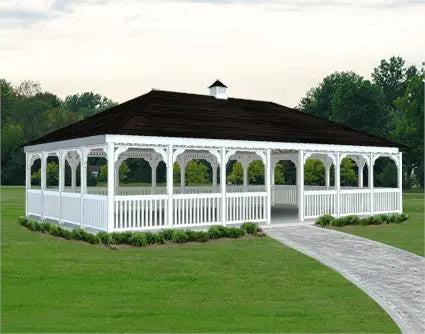 The image shows a large, white vinyl rectangular gazebo with a double roof and a cupola. The gazebo features a gabled roof, white railings, and a dark-colored roof. The gazebo is surrounded by green grass and trees, and there is a paved walkway leading up to it.