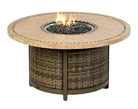 A round outdoor fire table with a woven base and a travertine stone tabletop. The table has a circular burner and blue flames are visible. There is a propane tank storage compartment at the base, and the table has four small wheels.