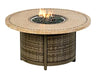 A round outdoor fire table with a woven base and a travertine stone tabletop. The table has a circular burner and blue flames are visible. There is a propane tank storage compartment at the base, and the table has four small wheels.