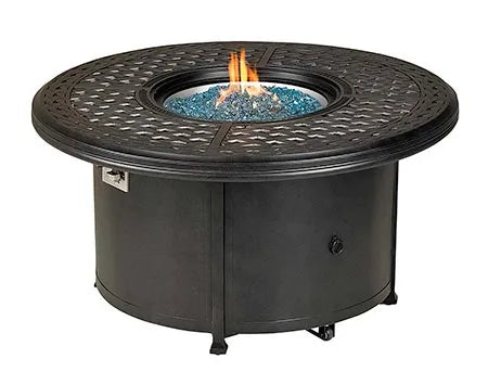 A round outdoor fire pit table with a black metal frame and a circular burner. Blue flames are visible through the decorative metal grate. There is a propane tank storage compartment at the base, and the table has four small wheels.