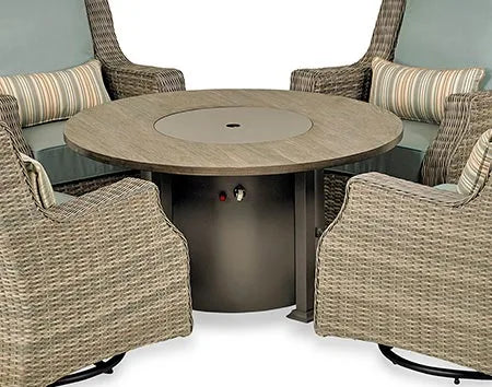 A close-up of a round fire pit table with a wooden top and a bronze-colored base. The table has a circular burner and a control panel with a knob and a switch. There is a metal cover over the burner, and the base has a storage compartment with a door.