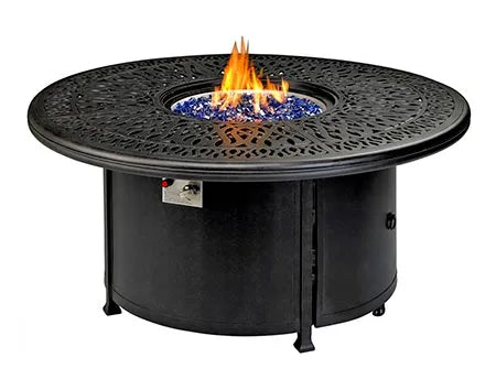  A round outdoor fire table with a black metal frame and a circular burner. Blue flames are visible through the decorative metal grate. There is a propane tank storage compartment at the base, and the table has four small wheels.