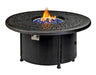  A round outdoor fire table with a black metal frame and a circular burner. Blue flames are visible through the decorative metal grate. There is a propane tank storage compartment at the base, and the table has four small wheels.