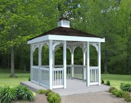 White vinyl gazebo with a double roof and a cupola. The gazebo has a rectangular shape with a gabled roof and white railings. The gazebo is surrounded by green trees and grass.