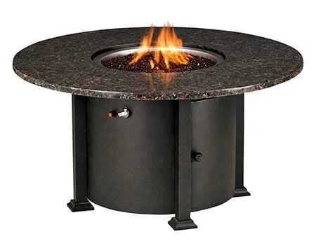 A round outdoor fire table with a granite top and a black metal base. The table has a circular burner and a control panel with a knob and a switch. A granite lid covers the burner, and a door on the side of the base reveals a storage compartment.