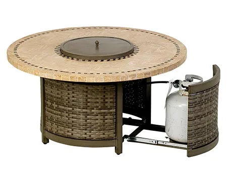 A round outdoor fire table with a woven base and a travertine stone tabletop. The table has a circular burner and blue flames are visible. There is a propane tank storage compartment at the base, and the table has four small wheels.