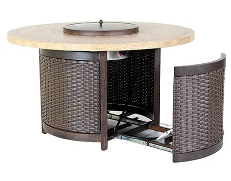 A round outdoor fire table with a woven base and a travertine stone tabletop. The table has a circular burner and blue flames are visible. There is a propane tank storage compartment at the base, and the table has four small wheels. The image shows a side view of the table with the storage compartment door open, revealing the propane tank inside.